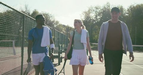 Walking tennis partners carrying rackets and towels on sunny outdoor court after match
