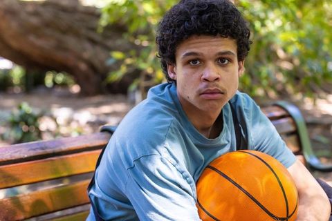 Contemplative african american teenager sitting on park bench holding basketball