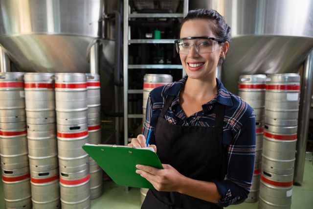 Female worker in a brewery holding a clipboard and smiling. She is wearing safety glasses and an apron, standing in front of large storage tanks and beer kegs. This image can be used for articles or advertisements related to the brewing industry, quality control, manufacturing processes, or workplace environments.