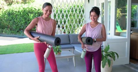Diverse women carrying yoga mats and water bottles on sunny patio preparing for outdoor yoga
