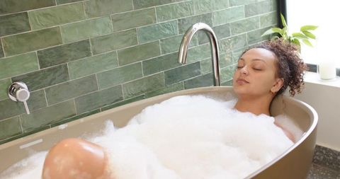 African american woman reclining in bubble bath in minimalist spa bathroom with green tile