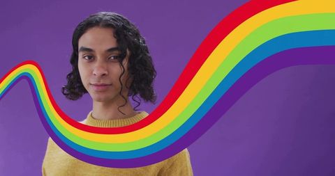 Nonbinary person wearing mustard sweater facing bold rainbow ribbon on purple background