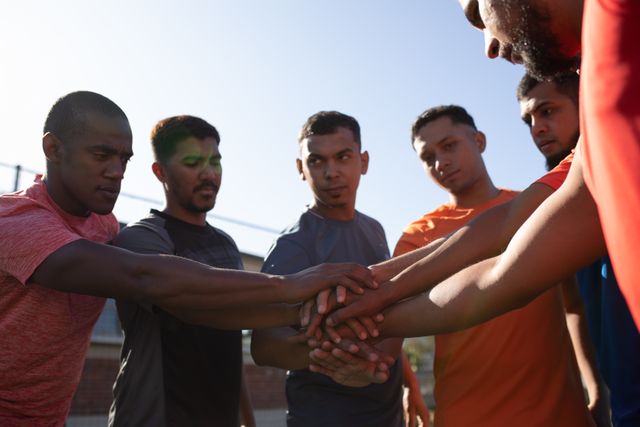Group of male football players from diverse backgrounds stacking hands in a show of unity and motivation before a game. Perfect for illustrating teamwork, sportsmanship, and the importance of unity in sports. Suitable for use in sports-related articles, team-building promotions, and fitness campaigns.