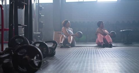 Two women performing seated medicine ball twists in backlit urban gym interior core workout