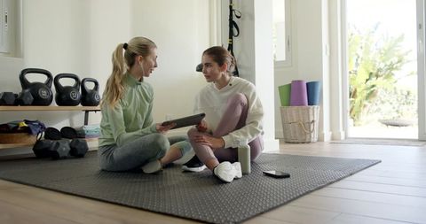 Two women planning home workout with tablet, kettlebells and yoga mats in bright fitness room