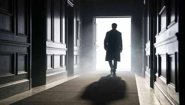 Silhouetted man walking toward bright doorway in fog-filled elegant corridor