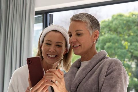 Mother and adult daughter sharing morning moment while checking smartphone in robes
