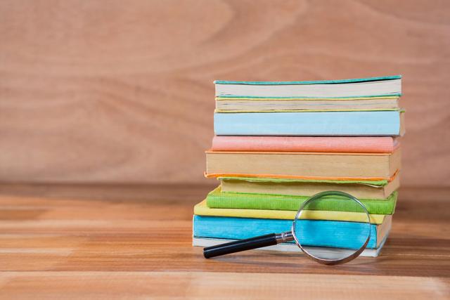 This image shows a magnifying glass placed next to a stack of colorful books on a wooden table. It is ideal for use in educational materials, library promotions, study guides, and research-related content. The vibrant colors of the books add a lively touch, making it suitable for both digital and print media focusing on learning and knowledge.
