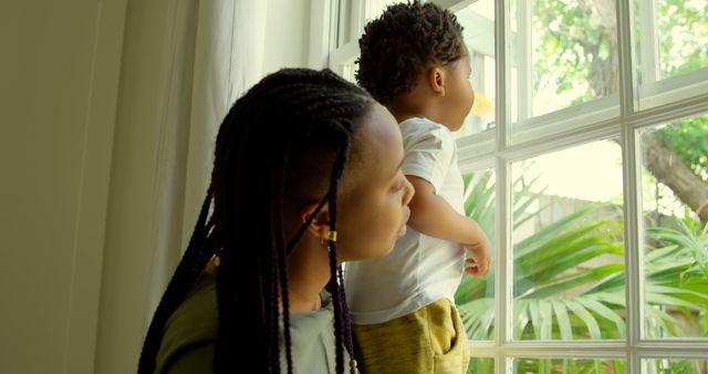 Side view of young black mother playing with his son on window sill in a comfortable home. They are looking outside through window 4k