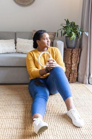 African american woman sitting on jute rug holding ring light and smartphone in cozy room