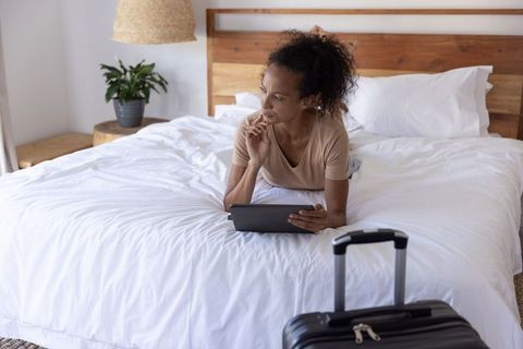 African american woman relaxing on bed using tablet with suitcase in modern hotel room
