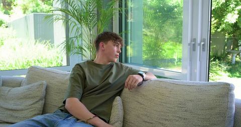 Teen boy relaxing on sectional sofa watching garden through large glass doors and windows