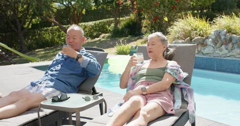 Senior asian couple relaxing on poolside loungers enjoying refreshing summer drinks