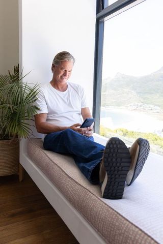 Mature man reclining on window bench using smartphone with sunlit coastal bay view