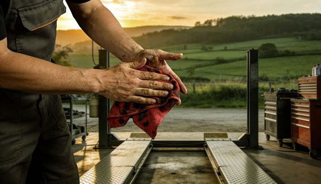 Mechanic wiping grease from hands with red rag at rural garage lift during golden sunset