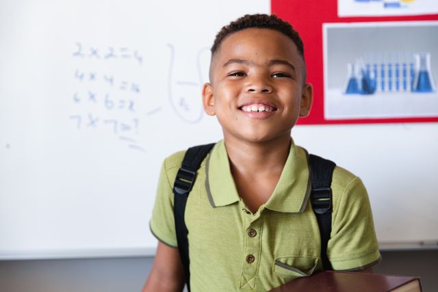 Young African American boy standing near whiteboard in classroom. He is wearing a green shirt and a backpack, smiling brightly. The board behind him shows mathematical equations and science diagrams. Ideal for use in educational materials, school brochures, websites about primary education, and articles focused on student life and learning.