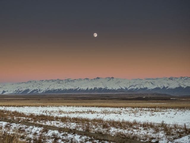 Full moon rising above snow-capped mountain range during dawn. Twilight sky creates serene ambiance over snowy, barren field. Ideal for backgrounds, travel brochures, environmental campaigns, and projects focused on natural beauty.