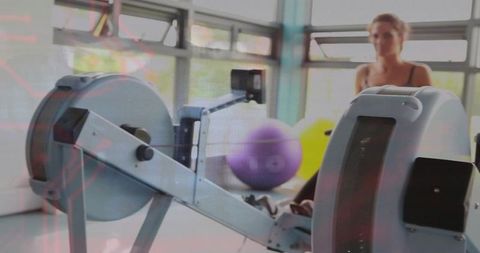 Female athlete rowing on ergometer in bright fitness studio with colorful stability balls