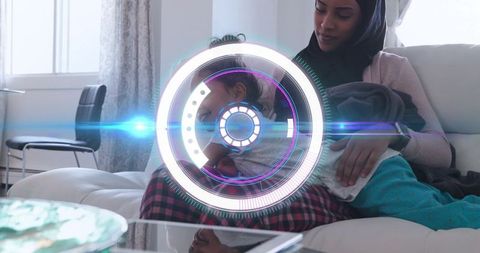 Cradling mother wearing hijab holding child on sofa with tablet and holographic interface