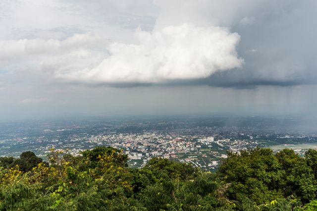 View of approaching rain clouds over a city with a mix of urban architecture and greenery below. Image can be used in articles or presentations about weather forecasts, climate change, urban life affected by weather, or scenic city views.