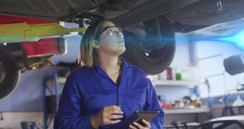 Female auto technician inspecting car undercarriage on lift with tablet and safety glasses