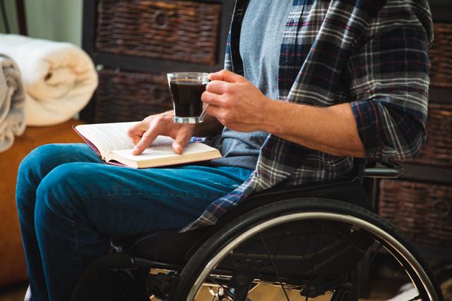 Disabled man in a wheelchair enjoying a quiet moment at home, reading a book and drinking coffee. Ideal for use in articles or advertisements related to disability awareness, home lifestyle, relaxation, and inclusive living.