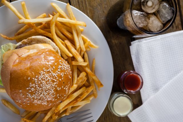 This image shows a close-up view of a classic fast food meal consisting of a burger with sesame bun, French fries, and a cold drink with ice on a wooden table. Condiments like ketchup and mayonnaise are also visible. Ideal for use in advertisements, menus, food blogs, or social media posts related to fast food, dining, or comfort food.