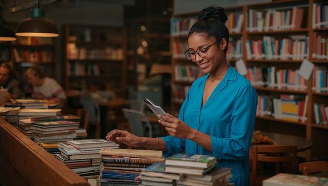 Smiling woman using smartphone at library counter amid stacked books and warm lighting