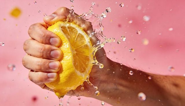 Squeezing lemon launching juice droplets on fresh pink background, closeup high-speed splash