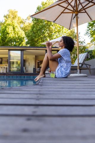 African american woman relaxing by modern backyard pool sipping cold drink under umbrella