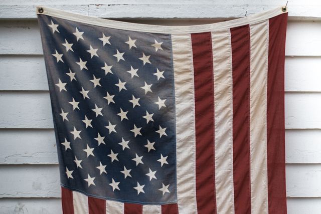 Vintage American flag with stars and stripes hanging against white wooden wall. Great for themes of patriotism, Americana, historical and vintage settings, and Fourth of July celebrations.