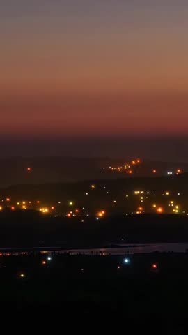 Vertical twilight time-lapse showing town lights rising across valley reflecting on river