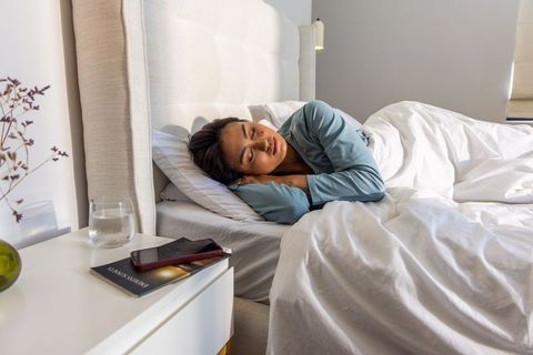 Asian woman in twenties resting in sunlit minimalist bedroom wearing blue pajamas