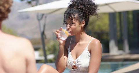 African american woman sipping lemon water poolside in striped one-piece swimsuit