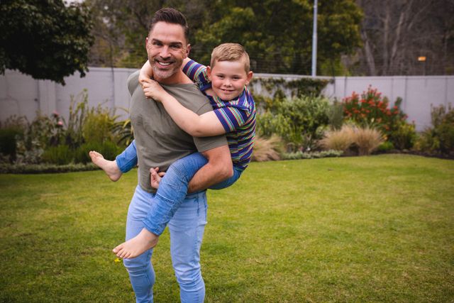 Father carrying his son on his back in a garden, both smiling and enjoying the moment. Ideal for use in family-oriented advertisements, parenting blogs, and lifestyle magazines. Highlights themes of fatherhood, love, and outdoor activities.