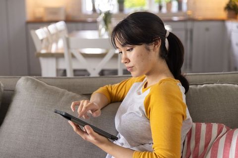 Asian woman relaxing on sofa using tablet for browsing in modern cozy home living room