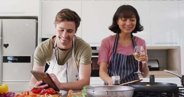 Image of happy diverse couple preparing meal together with tablet and wine. Love, relationship and spending quality time together concept.