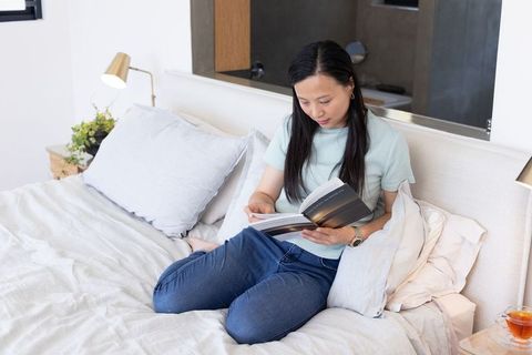 Mature african american woman reading paperback on bed in serene minimalist bedroom