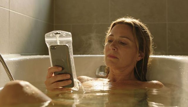 Middle-aged woman relaxing in steamy bathtub while using smartphone in waterproof pouch