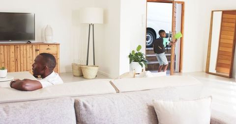 Father relaxing on couch while son entering home carrying watering can and backpack