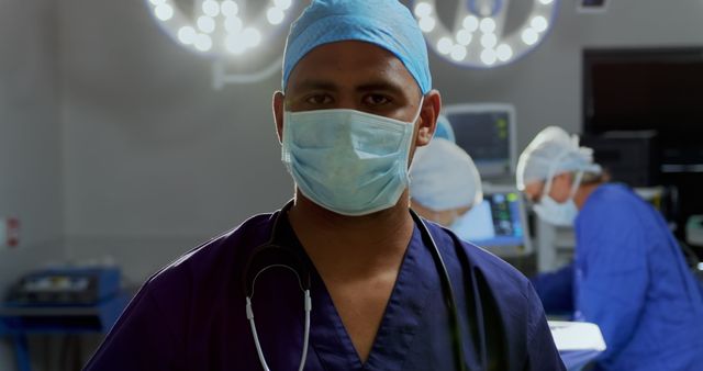 Front view of African-american male surgeon standing in the operation theater at hospital. Surgeons performing operation in the background