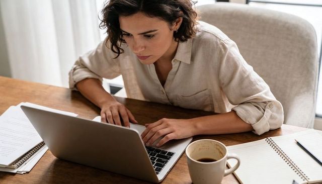 Focused woman typing on laptop at home desk with coffee and notebook for remote work