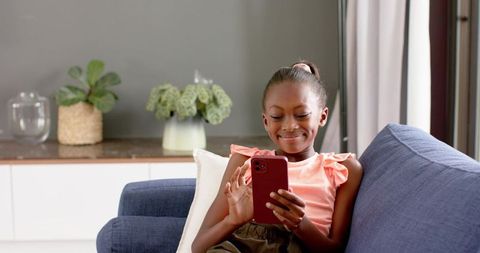 Preteen girl relaxing on blue sofa smiling while using red smartphone in cozy living room