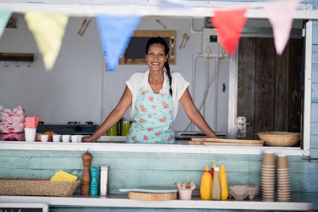 Waitress standing behind counter at outdoor food stall, smiling and wearing floral apron. Colorful bunting decorates the stall, creating a cheerful atmosphere. Ideal for use in articles about small businesses, food service, customer service, and outdoor events.