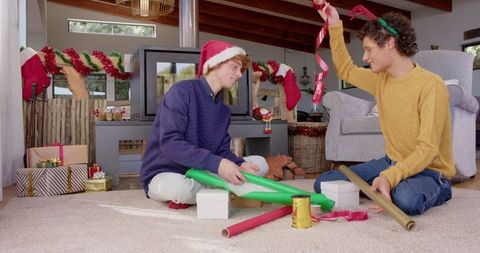 Gay male couple happily wrapping christmas presents by fireplace