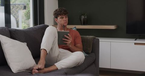 Young man lounging on modern sofa using tablet and stylus in minimalist living room