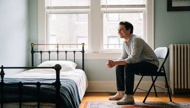 Young man sitting and smiling in minimalist sunlit bedroom with metal bedframe