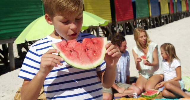 Boy in striped shirt enjoying juicy watermelon while sitting on the beach. Family is seen in the background having a picnic near colorful beach huts. Perfect for themes related to summer holidays, family vacations, healthy eating, and joyful childhood moments.