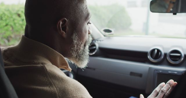 A senior African American man is driving a modern car through an urban environment. The man wears a light-colored sweater and has a gray beard. Great for websites and advertisements related to senior mobility, car safety, driver education for the elderly, or modern car features targeting mature drivers.