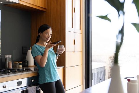 Asian woman speaking into smartphone while holding glass, leaning on wood kitchen cabinets